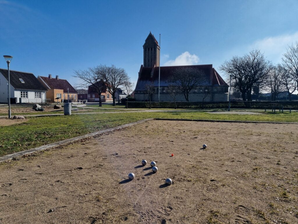 Pétanque balls on a gravel court with Gedser church and village houses behind under blue sky